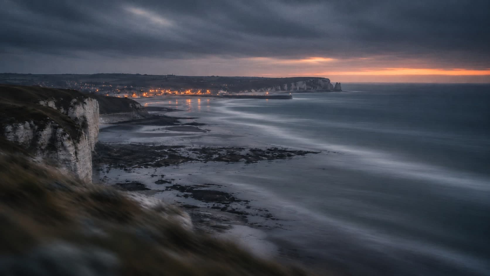 Chalk cliffs and a long-exposure sea at dusk, a seaside town lit in the distance