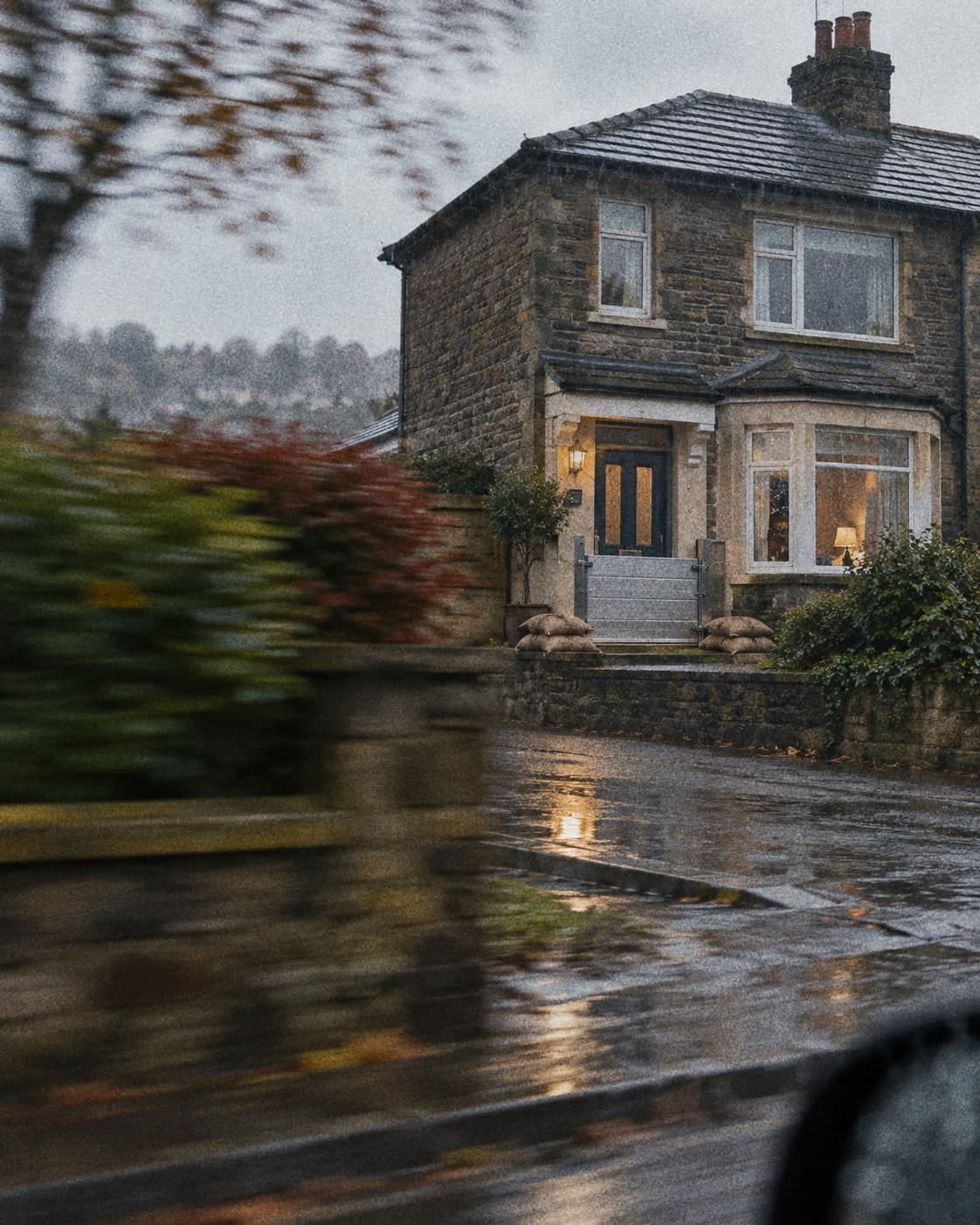 A stone semi-detached house with a flood gate and sandbags at the door, in the rain